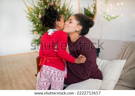 African American mother kissing daughter at home during  celebrating the Christmas holiday , New Year and exchanging gifts, Christmas tree in the background