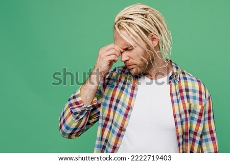 Young sad caucasian blond man with dreadlocks 20s he wear casual shirt keep eyes closed rub put hand on nose isolated on pastel plain light green background studio portrait. People lifestyle concept