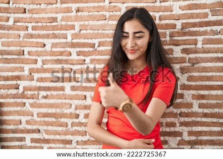 Young teenager girl standing over bricks wall looking proud, smiling doing thumbs up gesture to the side 