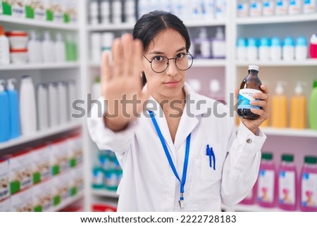 Young hispanic woman working at pharmacy drugstore holding syrup with open hand doing stop sign with serious and confident expression, defense gesture 