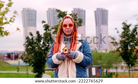 Positive young female in warm clothes and with long dyed dreadlocks holding small wooden house and looking at camera against blurred skyscrapers in city.