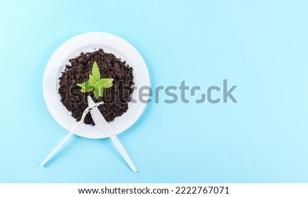 Plastic white plates with black earth and green sprout with crossed forks and knife on the left on a light blue background with copy space on the right, flat lay close-up. Ecology concept, 