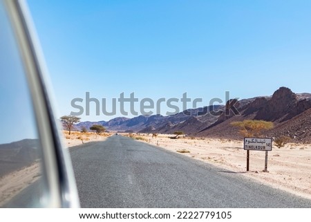 Tin El Koum, Algeria. Looking from rear 4X4 car window Tinelkoum desert road plaque with a dromedary on the reg, rocky mountains and blue sky background, Djanet district national road to Illizi city.