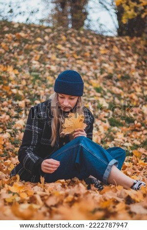 Smiling and happy brunette sitting in a pile of colourful leaves in a city park. Candid portrait of a young real woman in autumn clothes in the fall season.