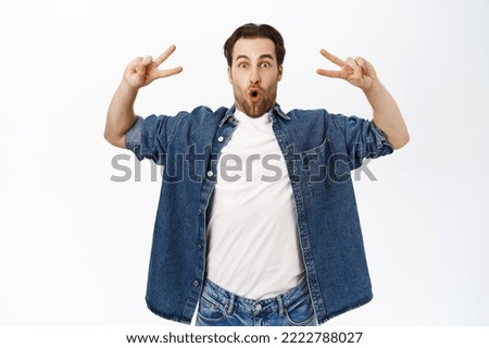 Positive handsome guy shows peace, v-signs and smiles happily, stands in casual clothes over white background. Copy space