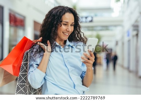 Happy and successful woman shopping for clothes in a supermarket store, Hispanic woman holding a smartphone reading online messages and browsing offers with discounts and sales Royalty-Free Stock Photo #2222789769