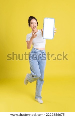 full body image of young Asian girl holding phone with cheerful face on yellow background Royalty-Free Stock Photo #2222856675
