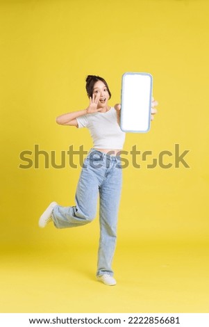 full body image of young Asian girl holding phone with cheerful face on yellow background
