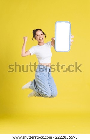 full body image of young Asian girl holding phone with cheerful face on yellow background Royalty-Free Stock Photo #2222856693