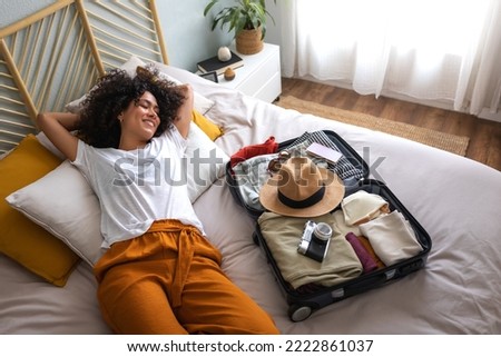 Happy woman lying down on bed next to open suitcase full of clothes, passport and hat, ready to go on vacation trip. Royalty-Free Stock Photo #2222861037