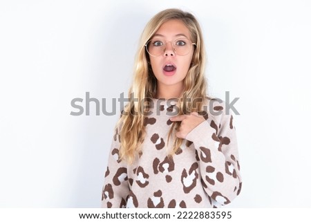 caucasian teen girl wearing animal print sweater over white wall being in stupor shocked, has astonished expression pointing at oneself with finger saying: Who me?
