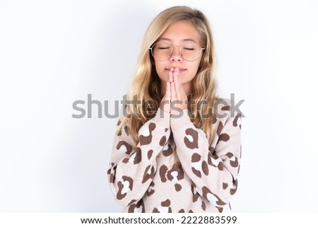 Indoor closeup of caucasian teen girl wearing animal print sweater over white wall practicing yoga and meditation, holding palms together in namaste, looking calm, relaxed and peaceful.