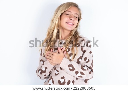 caucasian teen girl wearing animal print sweater over white wall closes eyes and keeps hands on chest near heart, expresses sincere emotions, being kind hearted and honest. Body language and real feel