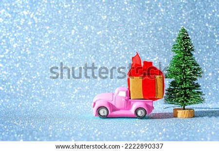Pink retro pickup truck with a large gift box parked by a toy Christmas tree against a sparkling background imitating snowflakes.