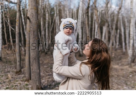 A beautiful young mother in a white dress and a beige coat with a baby in a spring birch grove