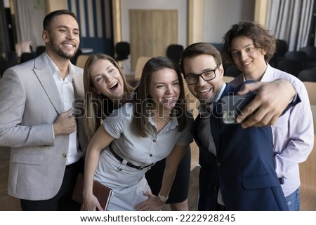 Happy corporate team, well-dressed company employees have fun at workplace, standing in modern office staring at smart phone screen make selfie picture. Friendly relations at work, modern tech, break