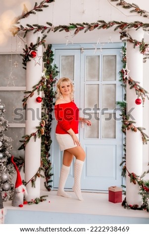 young woman in a red sweater stands on the porch of a house decorated for Christmas