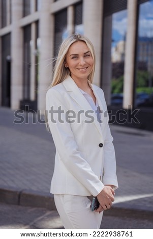 Vertical shoot of attractive young woman in white suit standing outdoors cute smiling at sunny day. Attractive caucasian blonde businesswoman toothy smiling satisfied by career and successful business