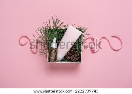 Beauty box of seasonal winter gifts on pink background. Cosmetic tube, serum and green fir branches with bump in cardboard packaging box with pink ribbon