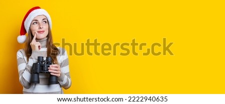 Pensive young woman in Santa Claus hat with binoculars on a yellow background. Banner.