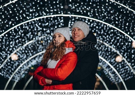 young happy loving couple hugging each other, looking somewhere together, smiling happily, christmas lights in background, caucasian boyfriend and girlfriend in red and black jackets, scarves and hats