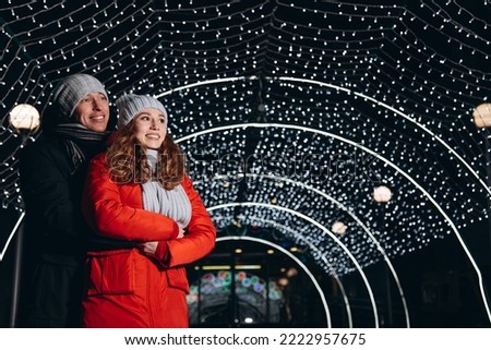 young happy loving couple hugging each other, looking somewhere together, smiling happily, christmas lights in background, caucasian guy and girl in red and black jackets, scarves and hats, copy space