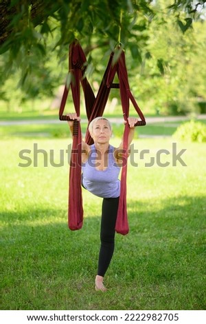 Athletic middle-aged woman in sportswear doing aerial yoga in a hammock outdoors, standing on one leg with her arms raised.The concept of aerial yoga, stretching,healthy lifestyle,mental and physical.