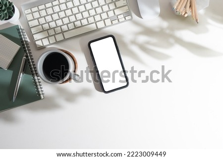 Flat lay mobile phone with empty screen, coffee cup, notepad and keyboard on white table. Top view with copy space.