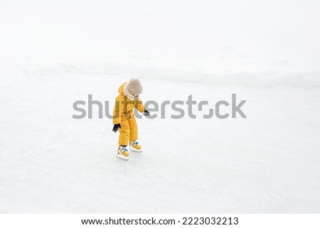 A small child in a warm yellow jumpsuit and a knitted hat is learning to skate by himself.  A lonely girl is skating alone on the lake