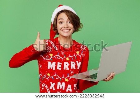 Merry young IT woman wear knitted xmas sweater Santa hat posing hold use work on laptop pc computer show thumb up isolated on plain pastel light green background. Happy New Year 2023 holiday concept