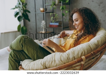 Side view young woman  wear casual clothes drink coffee read novel book sits in armchair stay at home flat rest relax spend free spare time in living room indoor grey wall