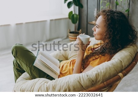 Sideways top view calm young woman wear t-shirt drink coffee read book sits in armchair stay at home flat rest relax spend free spare time in living room indoor grey wall Royalty-Free Stock Photo #2223032641