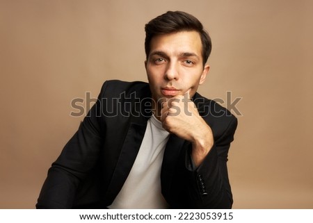 Portrait of handsome man wearing white t-shirt and black jacket, leaning on hand, posing on brown studio background, businessman looking at camera, ready to share his story of success