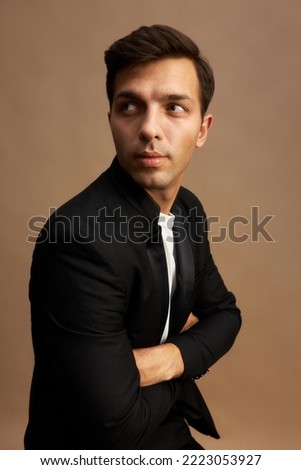 Vertical studio image of young guy with stylish haircut wearing jacket, sitting against brown background with folded arms, tuning head to the right, looking at something. Successful businesspeople