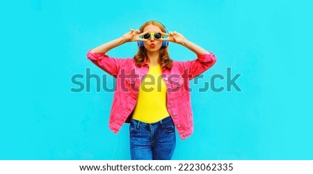 Portrait of stylish modern young woman in headphones listening to music blowing her lips sends sweet air kiss wearing pink jacket, yellow sunglasses on blue background