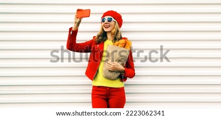 Portrait stylish happy smiling young woman taking selfie with smartphone holding grocery shopping paper bag with long white loaf bread wearing red beret, heart shaped sunglasses on white background