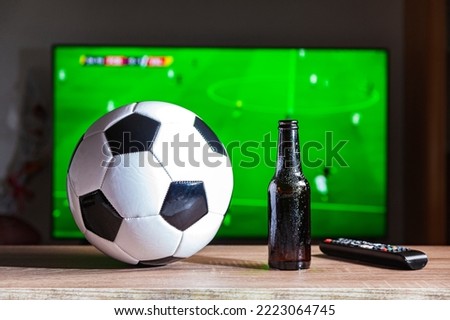 On a table is a soccer ball, a bottle of beer and a remote control. In the background, out of focus, there is a television set on which a football game is being broadcast.