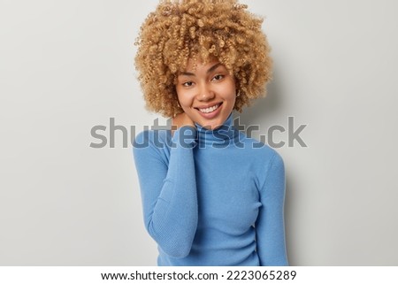Portrait of cheerful beautiful woman keeps hand on neck smiles pleasantly looks directly at camera feels delighted dressed in casual blue turtleneck isolated over grey background. Positive emotions