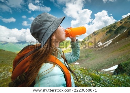 Profile photo of a young beautiful woman with long blond hair, dressed in a silver jacket, drinking from a yellow camping bottle, in the background a magnificent landscape of mountains and clouds