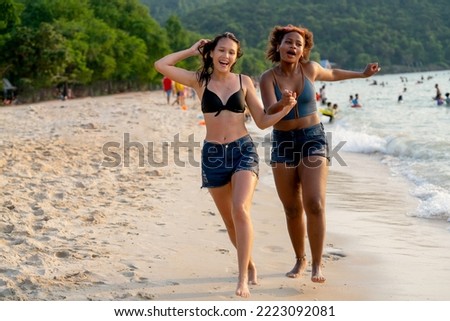 Two lovely teen girls hold hands and enjoy to run together on the beach with crowd of people in background during holiday or vacation.