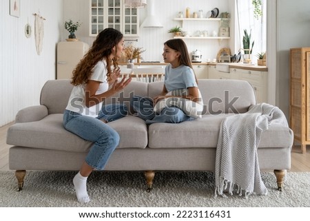 Tense teen girl holding pillow and looking at nervously gesturing mother sits on couch. Annoyed family quarrel between mother woman and daughter due to lack of time or money for mom to together walk. Royalty-Free Stock Photo #2223116431