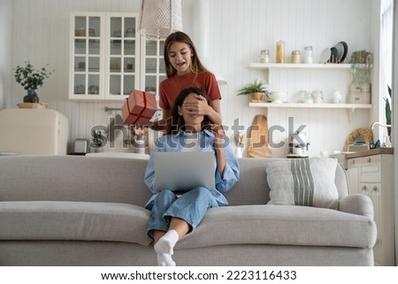 Teen girl daughter holding birthday gift congratulating happy young mother covering her eyes, congratulating with mothers day, kid making surprise for mom at home. Family and holidays concept