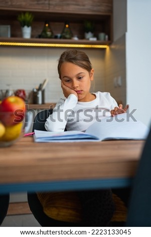 Upset little girl doing homework at table indoors, she's bored and wants to watch cartoons