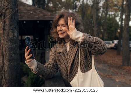Young smiling girl walking in autumn park taking selfie pictures using smartphone. Happy mood. Fashion style trend.