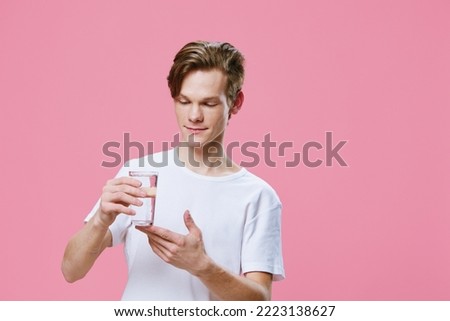 a cute, handsome guy in a white t-shirt stands on a pink background holding a glass of water in his hand and looks at him