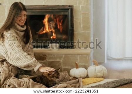 Happy woman caressing cute cat on cozy blanket at fireplace close up, autumn hygge. Stylish female in warm sweater together with kitty relaxing at fireplace in rustic farmhouse. Royalty-Free Stock Photo #2223241351
