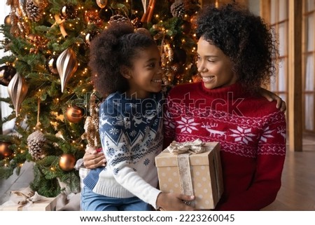 Smiling young African American mom and teen daughter near fir tree open Christmas gifts at home together. Happy biracial mother and kid celebrate New Year winter holidays unpack presents.