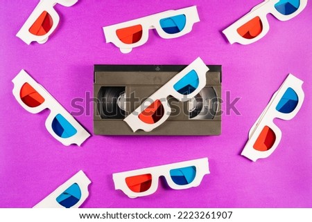 Old white paper 3d glasses with blue red lenses and black videotape vhs on purple background. Retro video cassette with magnetic tape and stereoscopic 3D cardboard glasses. Vintage background.