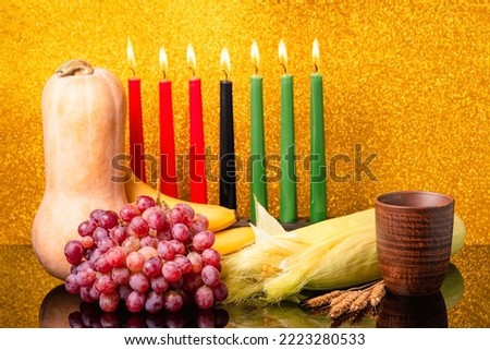 Kwanzaa holiday concept with decorate seven candles red, black and green, pumpkin, bowl, corns and fruit on light blur bokeh background with reflection