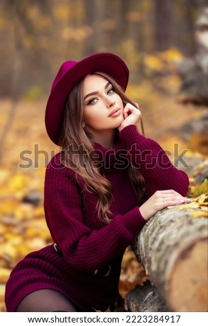 Beautiful girl in cosy knitted burgundy dress and hat sitting on nature with autumn background. Royalty-Free Stock Photo #2223284917